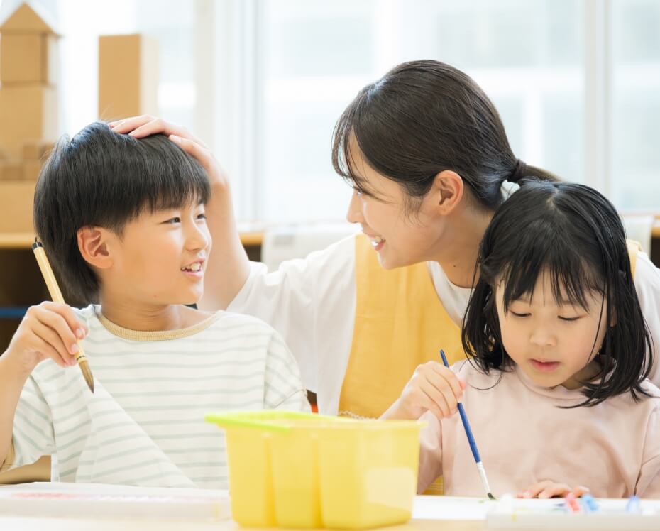 写真：工作作業中の児童たちと、そのうちの一人の頭を撫でて褒める保育士