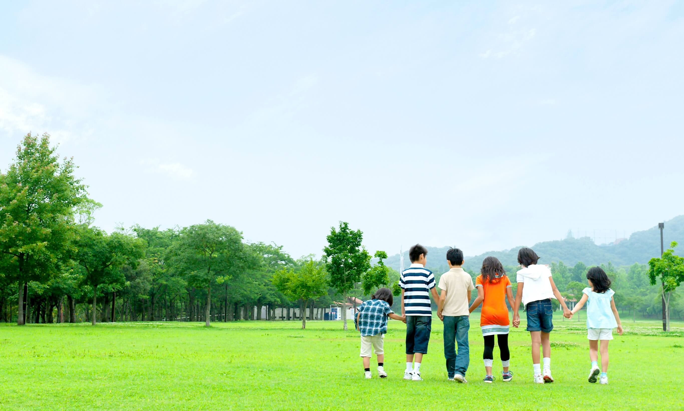 写真：周りは緑に囲まれた芝生が広がる広場にて、子どもたちが手を取り合いながら手前から奥に向かって歩いている。